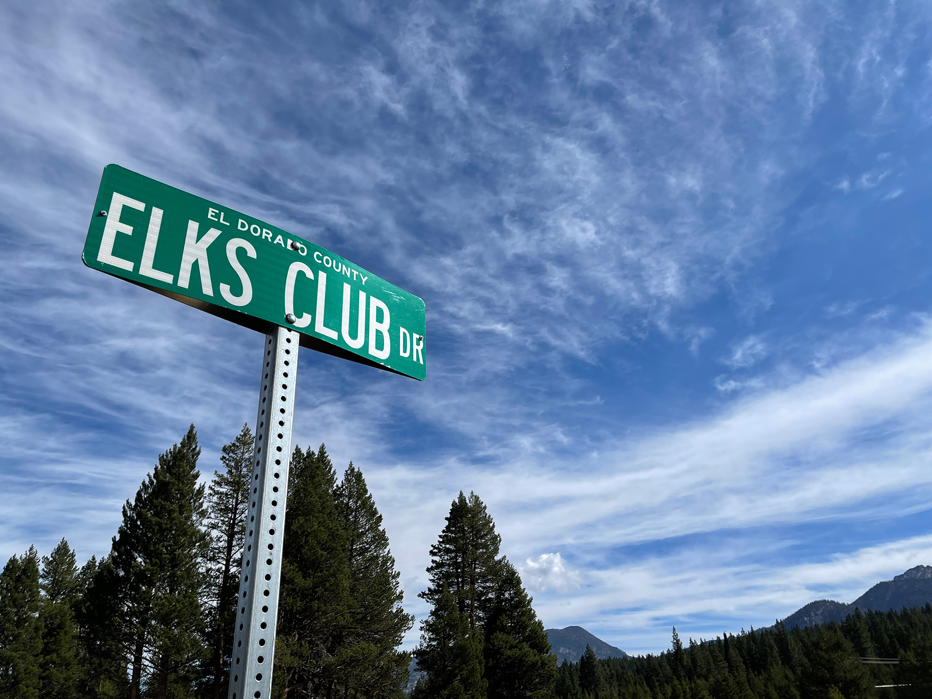 A street sign saying Elks Club Drive with partly cloudy skies above and mountains and trees in the background.